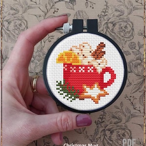 May include: A counted cross stitch pattern of a red mug with whipped cream, a cinnamon stick, and a star on a white background. The pattern is in a black embroidery hoop. The text "Christmas Mug" and "Counted cross stitch pattern" is below the hoop.