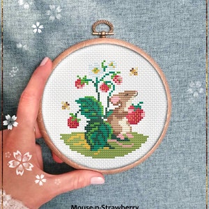 Puede incluir: Un patrón de punto de cruz contado que presenta un ratón marrón sosteniendo una fresa roja, con una planta de fresa verde y flores blancas en el fondo. El patrón está enmarcado en un aro de madera.