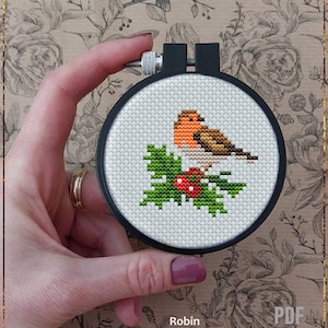 Può includere: Uno schema di punto croce contato che rappresenta un pettirosso appollaiato su un ramo di agrifoglio con bacche rosse. Lo schema è incorniciato in un cerchio da ricamo nero e il testo "Robin Counted cross stitch pattern" è sotto il cerchio.