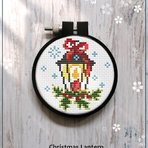 May include: A cross-stitch pattern of a Christmas lantern in a black embroidery hoop. The lantern is decorated with a red bow, holly, and snowflakes. The text "Christmas Lantern" and "Counted cross stitch pattern" are below the hoop.