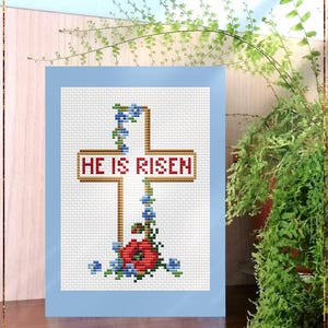 May include: A counted cross stitch pattern featuring a white cross with the words "HE IS RISEN" in red and gold lettering. The cross is adorned with blue flowers and a red flower at the base.