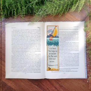 May include: Open book with a bookmark featuring a cross-stitch design. The bookmark has a sailboat image with the text "We have this hope as an anchor for the soul, firm and secure. Hebrews 6:19".