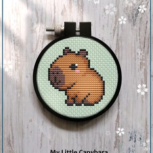 May include: A counted cross stitch pattern featuring a cute cartoon capybara. The pattern is in a round hoop with a light green background. The capybara is brown with a pink nose and is sitting on its haunches. The text "My Little Capybara" and "Counted cross stitch pattern" is below the hoop.