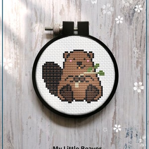 May include: Counted cross stitch pattern of a brown beaver holding a twig in its paws. The pattern is in a black embroidery hoop on a wooden background with white flowers.