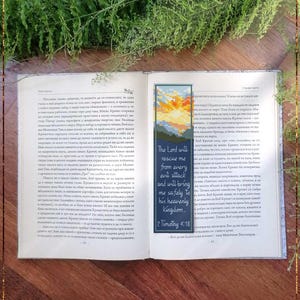 May include: A rectangular cross-stitch bookmark featuring a sunrise landscape in yellow, orange, and blue hues at the top, with the text "The Lord will rescue me from every evil attack and will bring me safely to his heavenly kingdom. 2 Timothy 4:18" below.