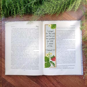 Op de afbeelding: Een rechthoekige kruissteek boekenlegger met een lichtblauwe achtergrond, omrand door groene bladeren, witte bloemen en rode aardbeien. De tekst luidt: I prayed for this child and the Lord has granted me what I asked of Him. 1 Samuel 1:27.
