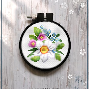 May include: A counted cross stitch pattern for a spring flower bouquet in a hoop. The flowers are white, pink, yellow, and blue. The pattern is labeled "Spring Flowers" and "Counted cross stitch pattern".