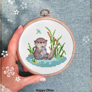 Op de afbeelding: Een borduurring met kruissteek met een bruine otter die een vis vasthoudt, omringd door groene riet en een blauwe libel. De tekst "Happy Otter" en "Counted cross stitch pattern" staan onder de afbeelding. De ring wordt door een hand vastgehouden.