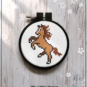 Puede incluir: Un patrón de punto de cruz contado para un caballo marrón con la nariz rosa, en un aro de bordado negro. El patrón se titula "My Little Horse" e incluye el texto "Counted cross stitch pattern".
