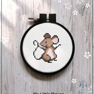 Könnte beinhalten: Eine Kreuzstich-Stickerei einer Cartoon-Maus in einem schwarzen Rahmen. Die Maus ist braun, hellbraun und rosa, mit schwarzen Details. Der Text "My Little Mouse" und "Counted cross stitch pattern" steht darunter.