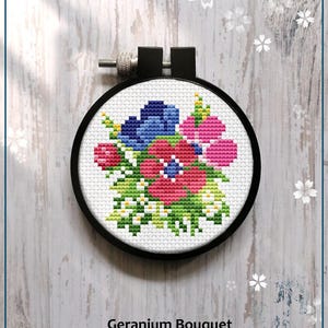 May include: A counted cross stitch pattern of a bouquet of pink, red, and blue flowers with green leaves and white flowers. The pattern is in a black embroidery hoop. The text "Geranium Bouquet" and "Counted cross stitch pattern" is below the hoop. The text "PDF" is in a white star shape to the right of the text.