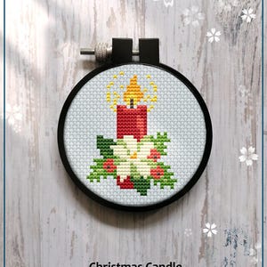 May include: A counted cross stitch pattern for a Christmas candle with a red candle, white flower, and green leaves. The pattern is in a black embroidery hoop on a white wooden background.