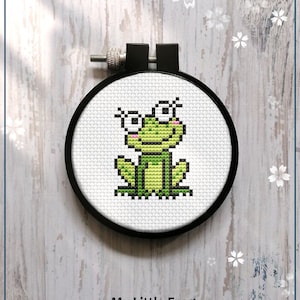 Può includere: Uno schema di punto croce contato per una rana verde con occhi grandi. Lo schema è in un cerchio da ricamo nero e il testo "My Little Frog" e "Counted cross stitch pattern" è sotto il cerchio. Il testo "PDF" è in un design di fiore bianco.