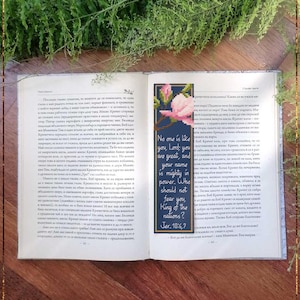 Puede incluir: Libro abierto con un marcapáginas de punto de cruz. El marcapáginas presenta un motivo floral y texto de Jeremías 10:6-7. El libro está sobre una superficie de madera con vegetación.