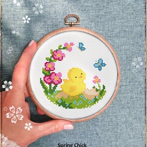 Op de afbeelding: Een geteld kruissteekpatroon van een geel kuiken dat in een nest van groen gras en roze bloemen zit met twee blauwe vlinders die erboven vliegen. Het patroon is ingelijst in een houten hoepel en de tekst "Spring Chick" en "Counted cross stitch pattern" staat onder de hoepel.