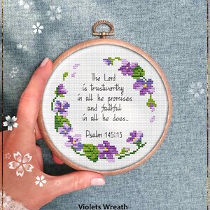May include: A round, wooden embroidery hoop holds a cross-stitch pattern. The design features a wreath of purple and green flowers surrounding the text "The Lord is trustworthy in all he promises and faithful in all he does. Psalm 145:13." The text "Violets Wreath" and "Counted cross stitch pattern" are below.