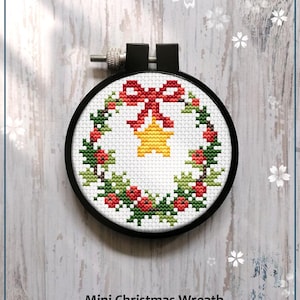 May include: A cross-stitch mini Christmas wreath design in a black embroidery hoop. The wreath features a red bow, a yellow star, green leaves, and red berries. The text "Mini Christmas Wreath" and "Counted cross stitch pattern" are below the wreath.
