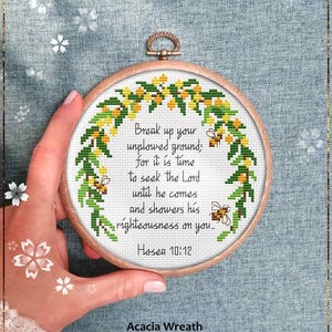 May include: A round, wooden embroidery hoop with a cross-stitch design. The design features a floral wreath with yellow and green leaves and bees, surrounding the text "Break up your unplowed ground... Hosea 10:12". The text "Acacia Wreath" and "Counted cross stitch pattern" are also visible.