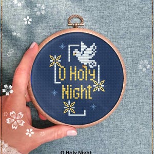 May include: A cross-stitch embroidery hoop with the words "O Holy Night" and a dove design. The text and dove are white and gold on a dark blue background. The wooden hoop is held by a hand.