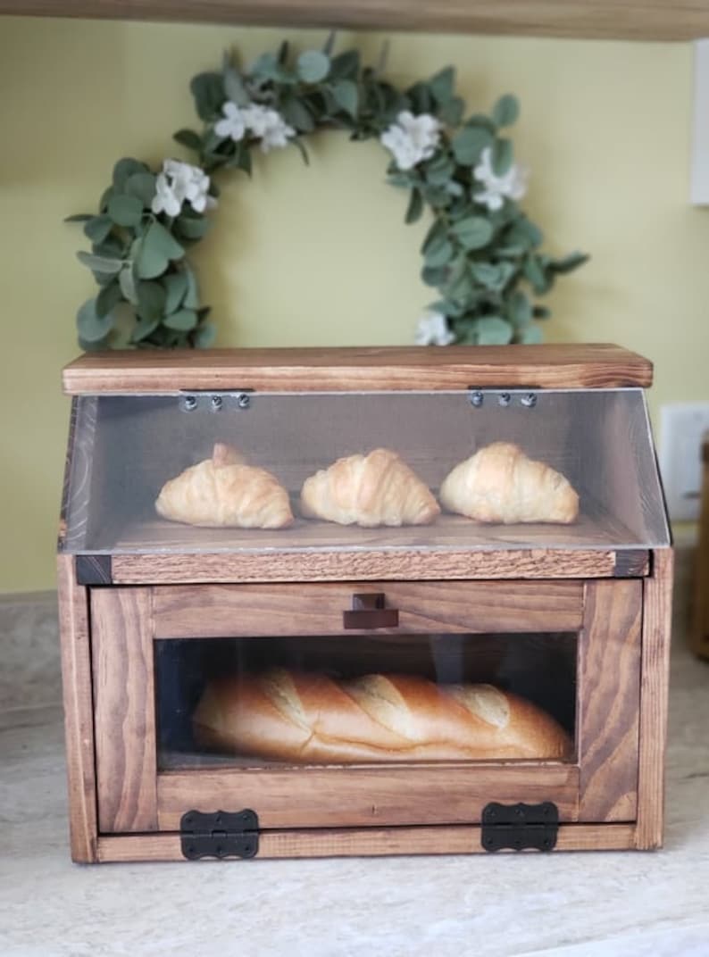 Puede incluir: Una caja de pan de madera con una puerta de vidrio y una tapa de vidrio. La caja est&aacute; llena de pan y croissants.
