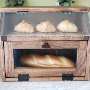 Puede incluir: Una caja de pan de madera con una puerta de vidrio y un caj&oacute;n. La repisa superior tiene tres croissants y la repisa inferior tiene una hogaza de pan.