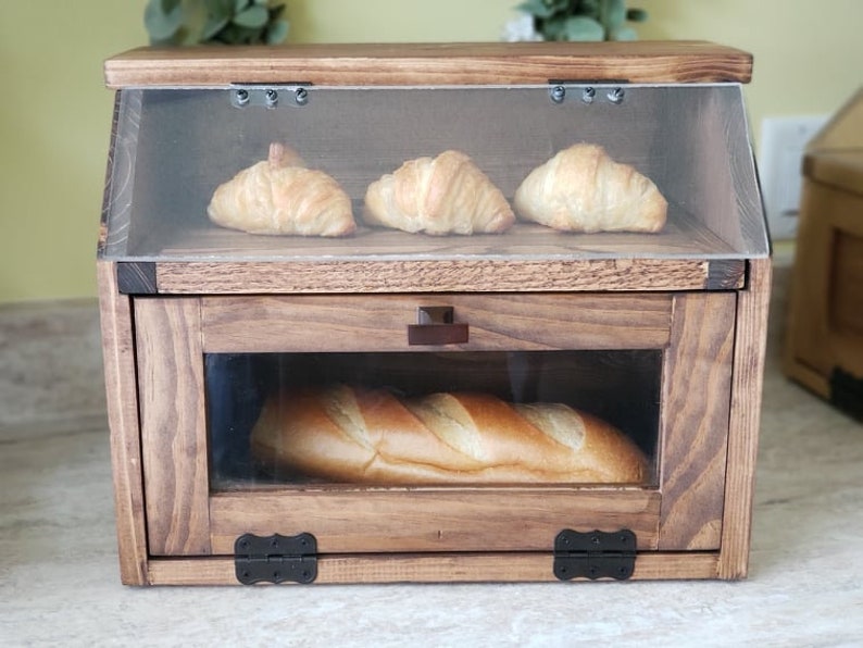 Puede incluir: Una caja de pan de madera con una puerta de vidrio y un caj&oacute;n. La repisa superior tiene tres croissants y la repisa inferior tiene una hogaza de pan.