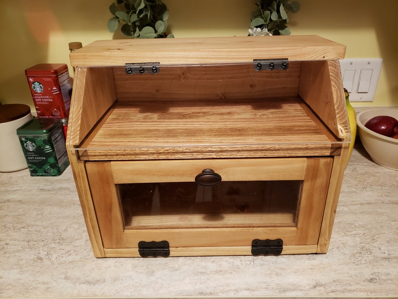 Puede incluir: Una caja de pan de madera con una puerta de vidrio y una tapa abatible. La caja tiene una perilla marr&oacute;n oscuro y bisagras negras.
