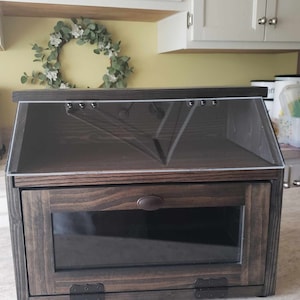 Puede incluir: Una caja de pan de madera marr&oacute;n oscuro con una tapa de vidrio transparente y una puerta de vidrio en la parte delantera. La caja tiene una manija y bisagras de metal.