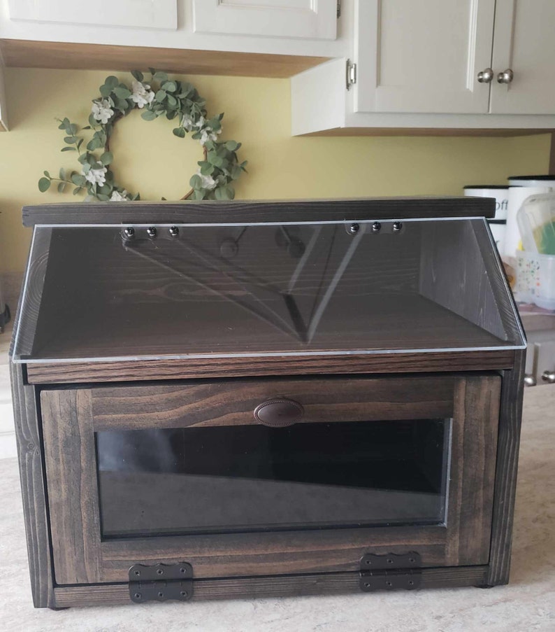 Puede incluir: Una caja de pan de madera marr&oacute;n oscuro con una tapa de vidrio transparente y una puerta de vidrio en la parte delantera. La caja tiene una manija y bisagras de metal.