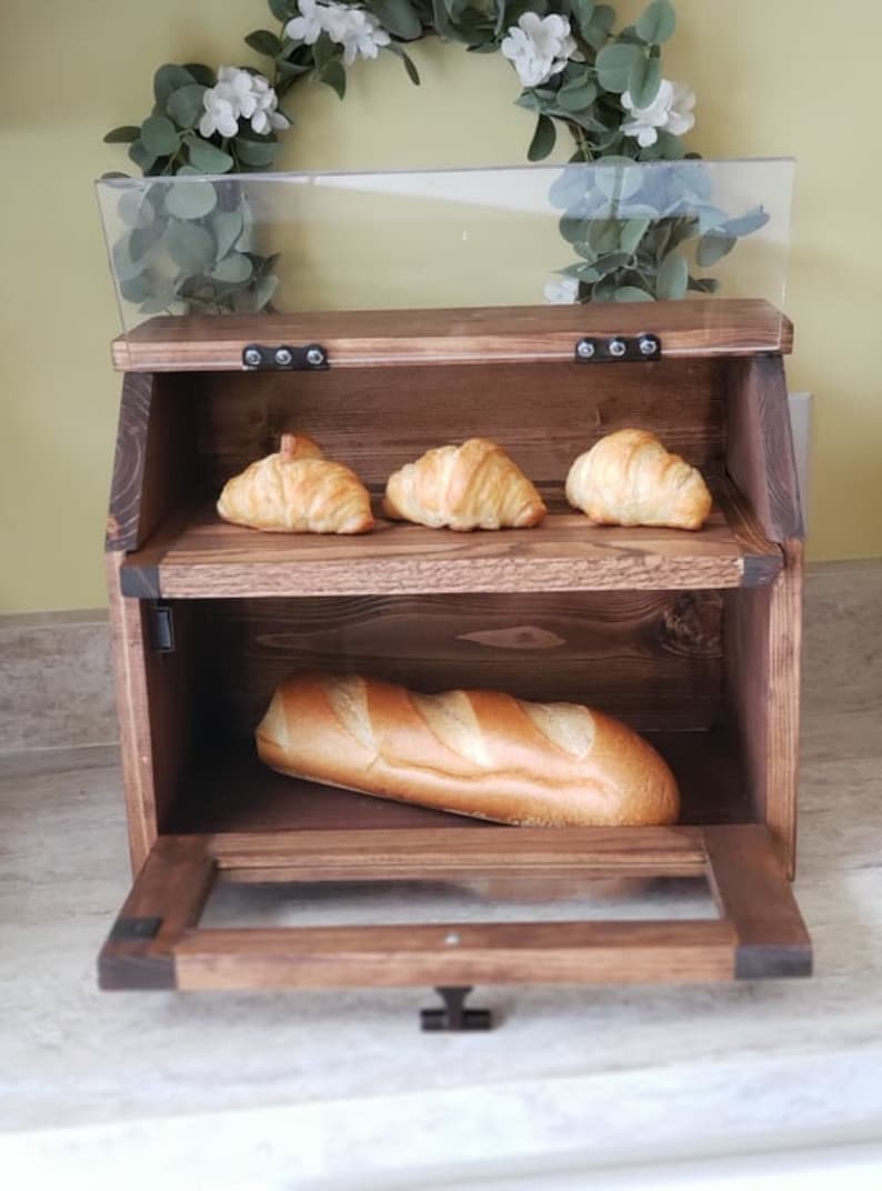 Puede incluir: Una caja de pan de madera con una puerta de vidrio y dos estantes. El estante superior tiene tres croissants y el estante inferior tiene una baguette.