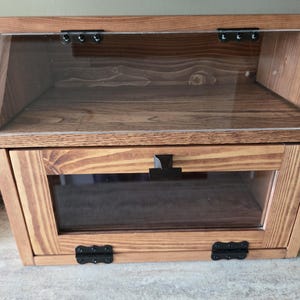 May include: A wooden bread box with a natural brown wood grain finish, featuring a clear acrylic slanted top lid and a front-opening door with a clear window panel. The unit includes black metal hinges and a black handle on the front door.