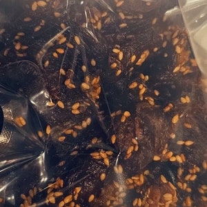 May include: A clear plastic bag filled with dark brown, chewy, sesame seed-covered beef jerky.