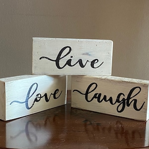 May include: Three white wooden blocks with black calligraphy. The blocks read "live", "love", and "laugh".