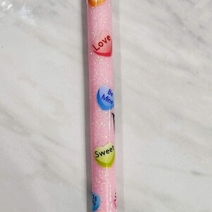 Puede incluir: Un bol&iacute;grafo rosa brillante con gr&aacute;ficos de caramelos en forma de coraz&oacute;n que dicen "Kiss", "Love", "Be Mine", "Sweet" y "Kiss You". El bol&iacute;grafo tiene una punta plateada y una punta de tinta negra.