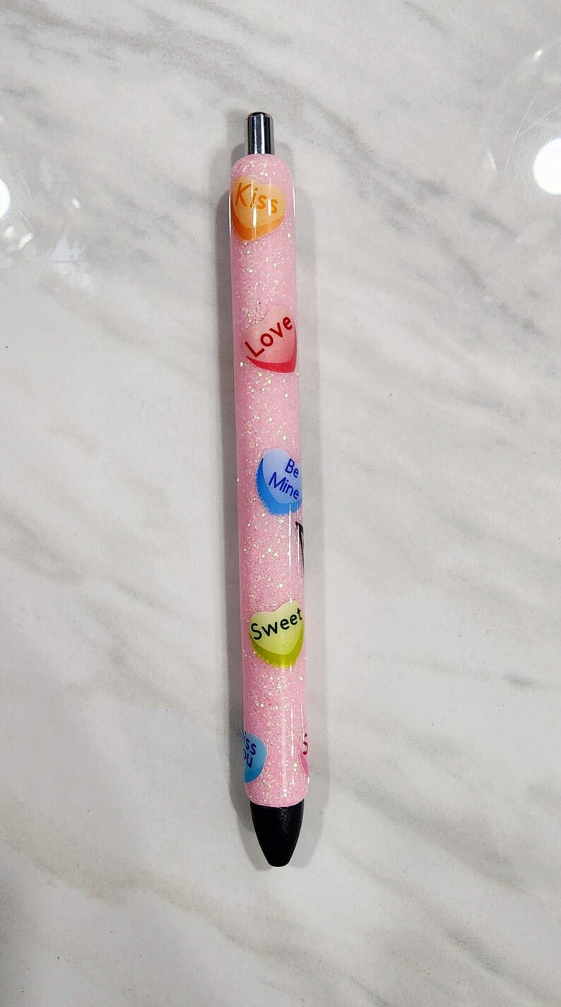 Puede incluir: Un bol&iacute;grafo rosa brillante con gr&aacute;ficos de caramelos en forma de coraz&oacute;n que dicen "Kiss", "Love", "Be Mine", "Sweet" y "Kiss You". El bol&iacute;grafo tiene una punta plateada y una punta de tinta negra.