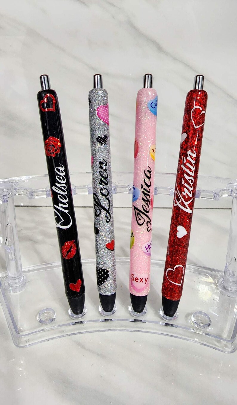 Puede incluir: Cuatro bol&iacute;grafos personalizados en un soporte transparente. Los bol&iacute;grafos son negros, plateados, rosas y rojos, con purpurina y dise&ntilde;os de corazones. Cada bol&iacute;grafo tiene un nombre: Chelsea, Loren, Jessica y Kristin.