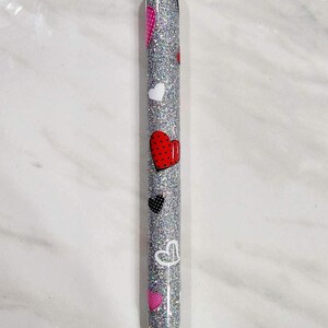Puede incluir: Un bol&iacute;grafo plateado con purpurina decorado con varios dise&ntilde;os de corazones en rojo, blanco, negro y rosa. El bol&iacute;grafo tiene una punta negra y detalles plateados.