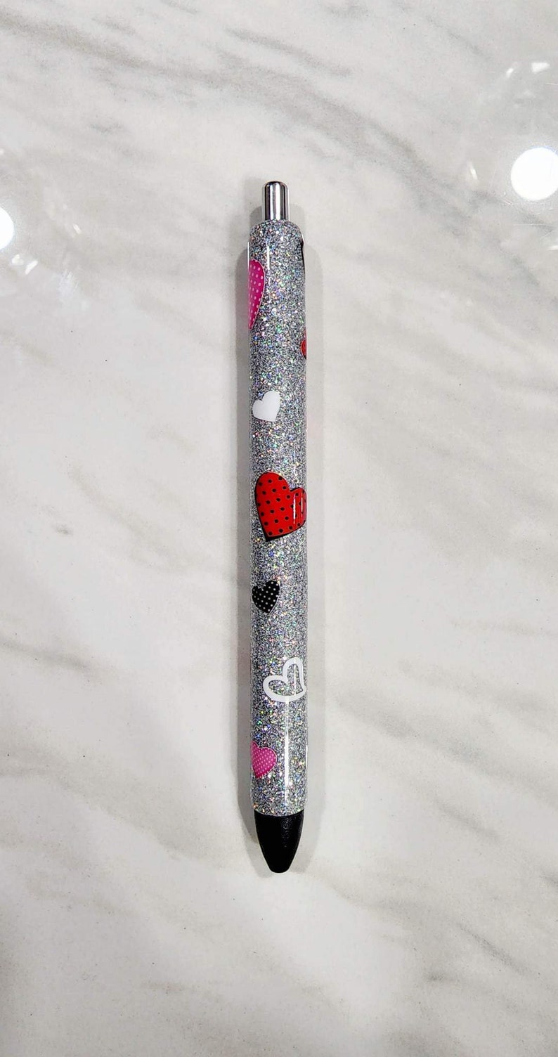Puede incluir: Un bol&iacute;grafo plateado con purpurina decorado con varios dise&ntilde;os de corazones en rojo, blanco, negro y rosa. El bol&iacute;grafo tiene una punta negra y detalles plateados.