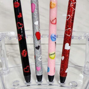 Puede incluir: Cuatro bol&iacute;grafos decorativos con temas de San Valent&iacute;n. Los bol&iacute;grafos tienen barriles brillantes en negro, plateado, rosa y rojo. Cada bol&iacute;grafo est&aacute; decorado con corazones, labios y dise&ntilde;os de corazones de caramelo. Los bol&iacute;grafos se exhiben en un soporte de pl&aacute;stico transparente.