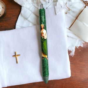 May include: A green glitter pen featuring an image of a religious figure. The pen has a silver tip and a dark green grip. A small gold cross and white fabric are also visible.