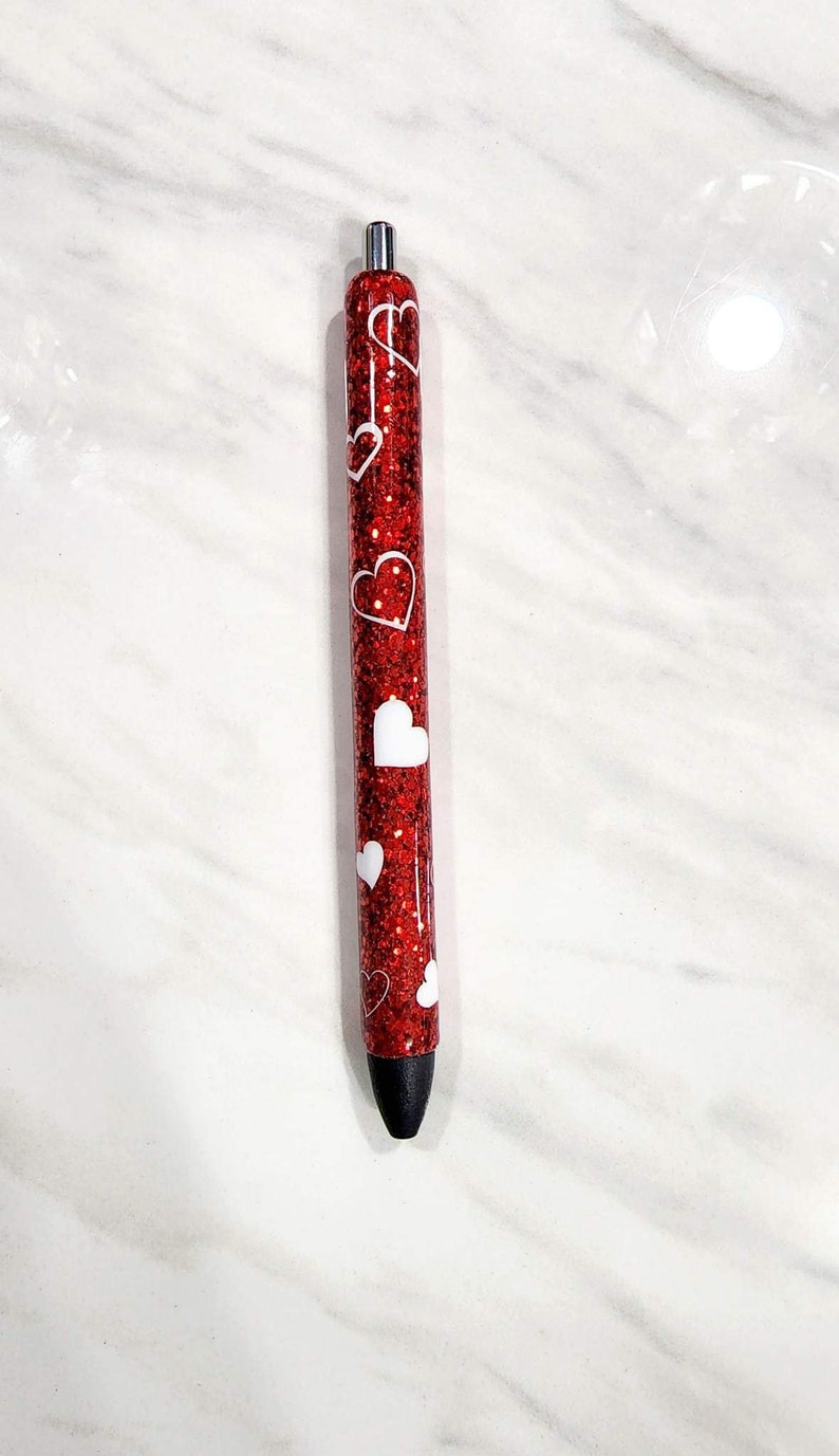 Puede incluir: Un bol&iacute;grafo rojo brillante con dise&ntilde;os de corazones blancos. El bol&iacute;grafo tiene una punta negra y detalles plateados. Es un instrumento de escritura y probablemente se utiliza para escribir diarios, tomar notas o escribir de forma creativa.