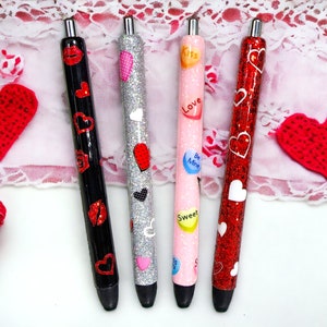 Puede incluir: Cuatro bol&iacute;grafos decorativos con temas de San Valent&iacute;n. Un bol&iacute;grafo negro presenta labios y corazones rojos, un bol&iacute;grafo plateado tiene corazones, un bol&iacute;grafo rosa tiene corazones de caramelo con mensajes y un bol&iacute;grafo rojo con purpurina tiene corazones blancos. Los bol&iacute;grafos est&aacute;n sobre una superficie blanca con decoraciones de coraz&oacute;n rojo.