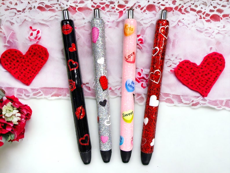 Puede incluir: Cuatro bol&iacute;grafos decorativos con temas de San Valent&iacute;n. Un bol&iacute;grafo negro presenta labios y corazones rojos, un bol&iacute;grafo plateado tiene corazones, un bol&iacute;grafo rosa tiene corazones de caramelo con mensajes y un bol&iacute;grafo rojo con purpurina tiene corazones blancos. Los bol&iacute;grafos est&aacute;n sobre una superficie blanca con decoraciones de coraz&oacute;n rojo.