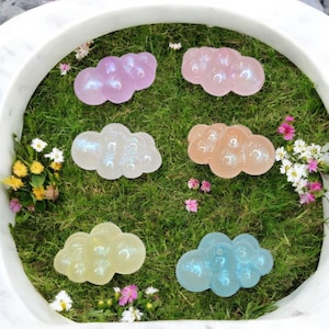May include: Six cloud-shaped soaps in various colors, including pink, purple, yellow, and blue, are arranged on a bed of green grass. The soaps have a pearlescent sheen and are surrounded by small flowers.