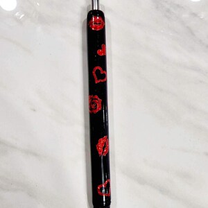 Puede incluir: Bol&iacute;grafo negro decorado con labios, corazones y una rosa de purpurina roja. El bol&iacute;grafo tiene una punta de metal plateado y est&aacute; sobre un fondo de m&aacute;rmol blanco. Un elegante accesorio de escritura.