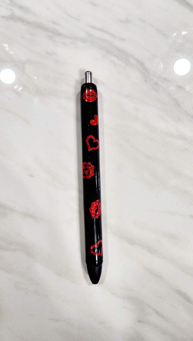 Puede incluir: Bol&iacute;grafo negro decorado con labios, corazones y una rosa de purpurina roja. El bol&iacute;grafo tiene una punta de metal plateado y est&aacute; sobre un fondo de m&aacute;rmol blanco. Un elegante accesorio de escritura.