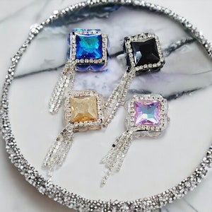May include: Four pairs of square-shaped earrings with different colored center stones: blue, black, gold, and iridescent purple. Each earring has a silver-colored frame and a dangling chain of small, clear crystals. The earrings are displayed on a white, marble-patterned surface.