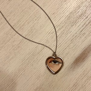 May include: A silver chain necklace with a heart-shaped pendant featuring a painted eye.