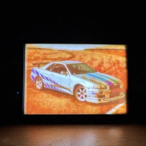 Op de afbeelding: Een lichtblauwe sportwagen met kleurrijke strepen wordt op een scherm weergegeven. De auto staat op een baan met een woestijnachtige achtergrond. De afbeelding wordt omlijst door een zwarte rand.