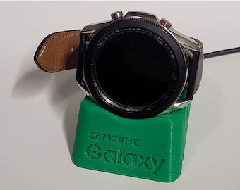 May include: A green charging stand for a Samsung Galaxy smartwatch. The stand has the words "Samsung Galaxy" printed on it. The smartwatch is on the stand and is connected to a charging cable.